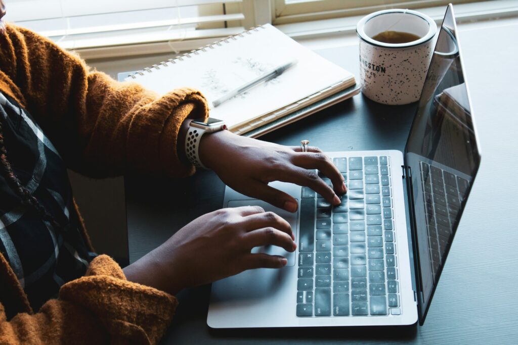 Hände tippen auf einem Laptop, daneben Notizbuch und Kaffeetasse