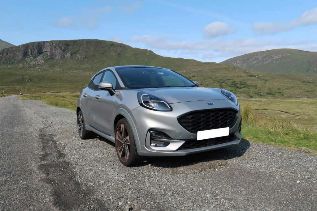 Ford Puma argenté sur une place de gravier au bord d’une route de campagne devant des collines vertes