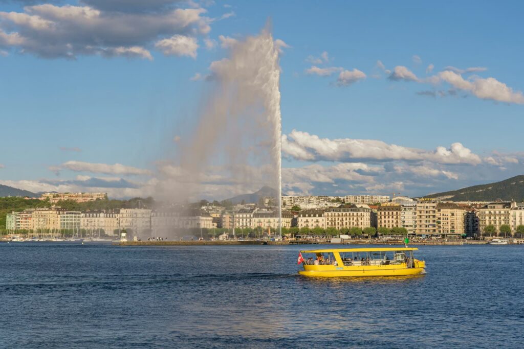 Il Jet d’eau nella rada di Ginevra, in primo piano una barca gialla delle Mouettes, sullo sfondo le case sul lungolago