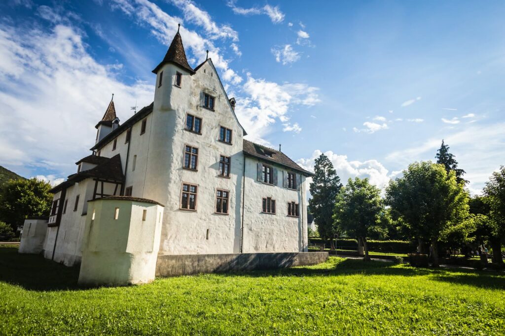 Le château de Pratteln aux murs blancs et à la tour d’angle pointue, sur une prairie sous un ciel bleu