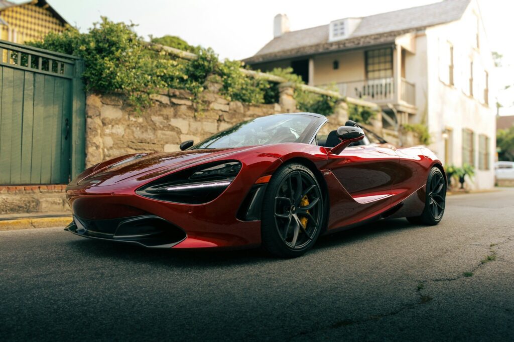 Roter McLaren 720S Spider mit offenem Dach von schräg vorne auf einer Gasse vor einer Bruchsteinmauer und einem hellen Haus