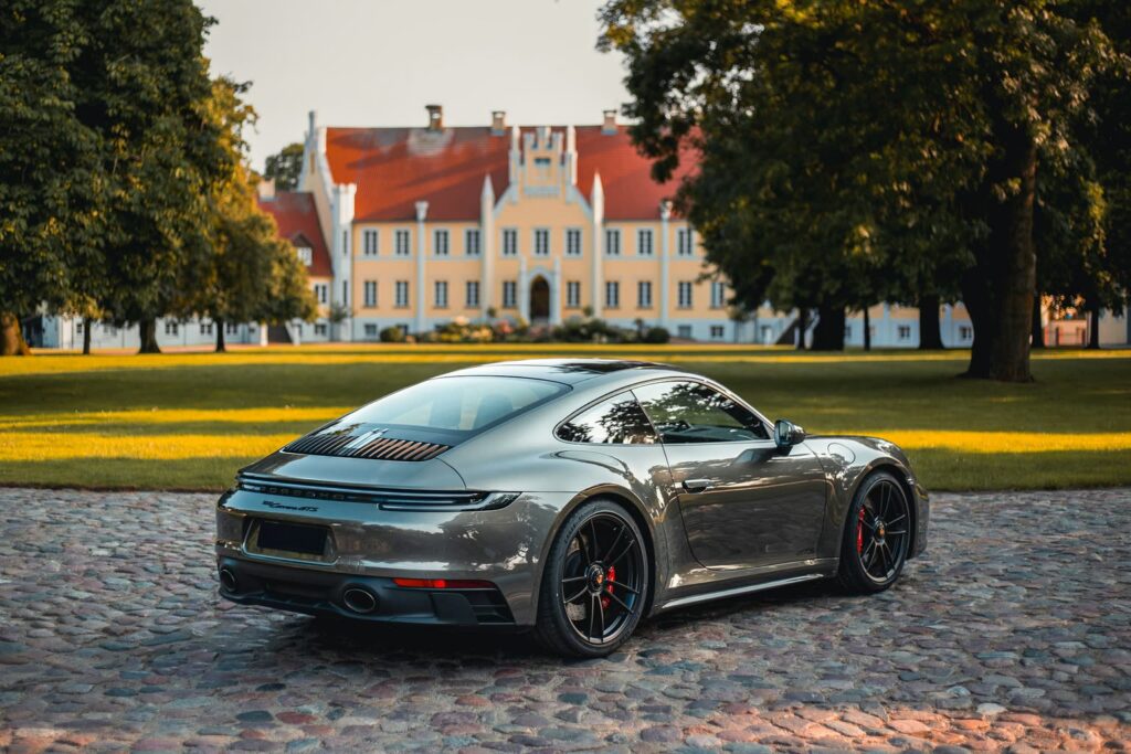 Grauer Porsche 911 Carrera 4 GTS von hinten, auf Kopfsteinpflaster vor einem Herrenhaus mit alten Bäumen