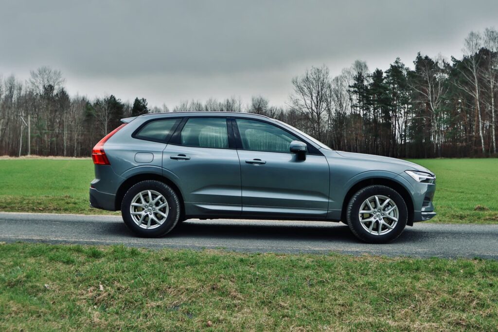 Volvo XC60 gris vu de côté sur une route étroite entre des prés, avec une lisière de forêt en arrière-plan